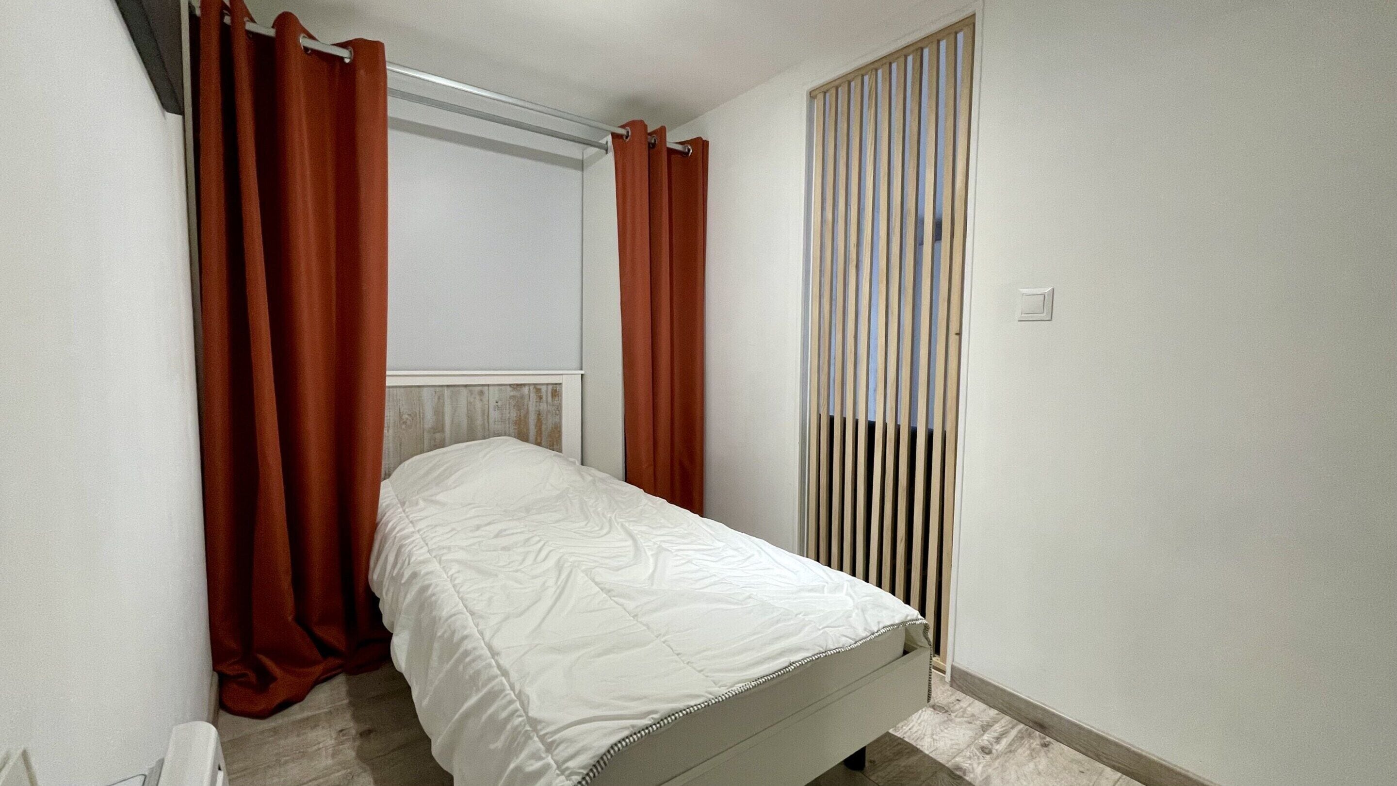 Um quarto pequeno e estreito, com uma cama de solteiro, uma janela com persianas verticais e um radiador na parede.