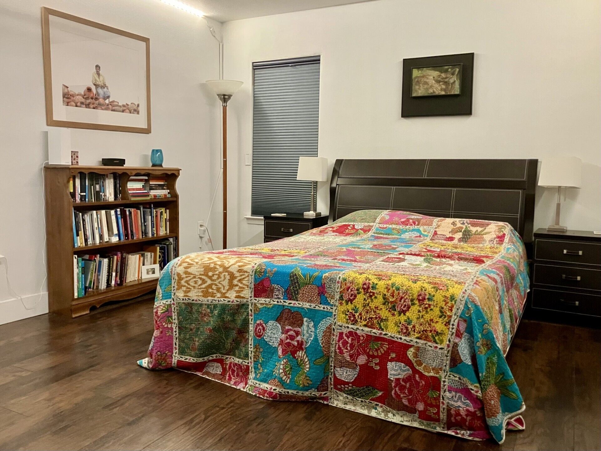 A bedroom with a large bed, a bookshelf, a lamp, and a window with blinds.
