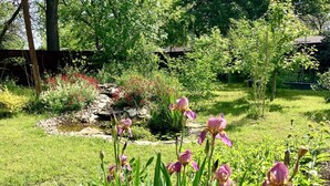 Ein Garten mit blühenden rosa Schwertlilien, einem kleinen Teich und verschiedenen Sträuchern und Bäumen.