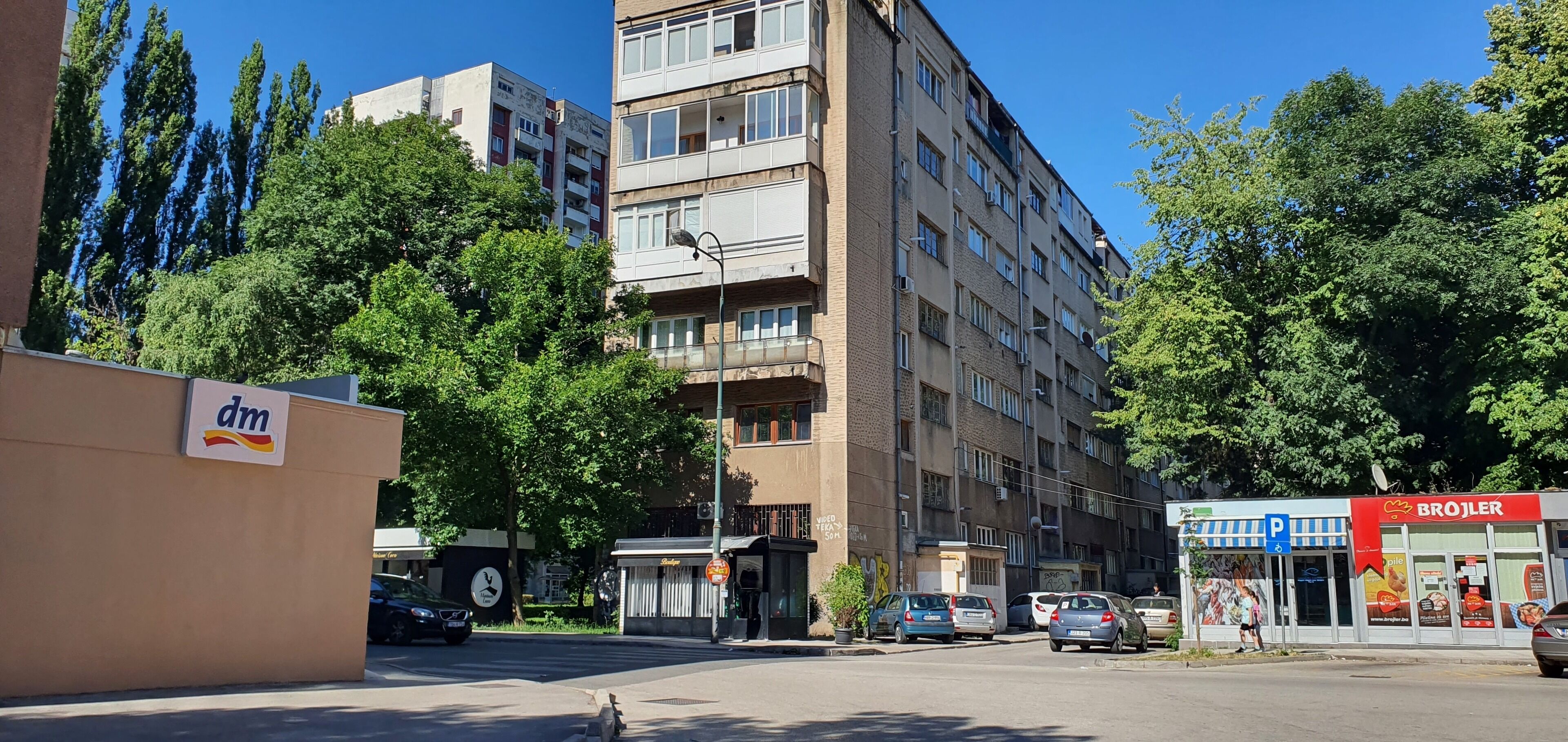 Een meerlaags appartementencomplex met balkons, een parkeerplaats en een winkel met een rood bord.