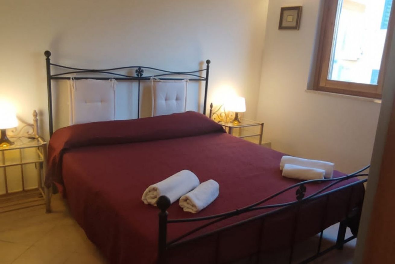 Un lit bien fait, recouvert d’une couverture rouge, avec deux serviettes enroulées. Il y a deux tables de chevet avec des lampes, et une fenêtre avec des rideaux.