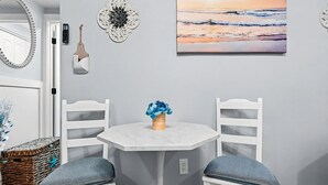 Ein Essbereich mit einem weißen Tisch und Stühlen, einer Vase mit blauen Blumen, einem Wanddekor und einem Gemälde, das einen Sonnenuntergang über dem Meer zeigt.