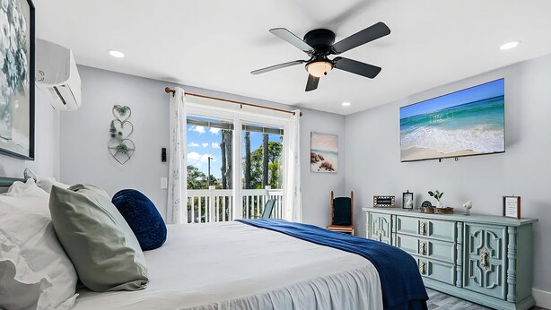 Ein Schlafzimmer mit einem Bett, einem Deckenventilator, einem großen Fenster mit Blick auf den Strand und einem Fernseher an der Wand.