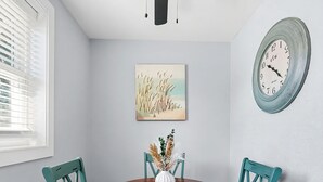 Un comedor con una mesa redonda, sillas color turquesa, un jarrón blanco con plantas secas, un reloj de pared y un cuadro.