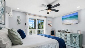Un dormitorio con cama, ventilador de techo, ventana grande con vista a la playa y televisor en la pared.