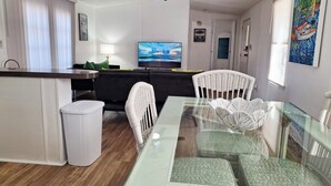 Una sala de estar moderna con una mesa de comedor de vidrio, sillas de mimbre blanco, un sofá negro y un televisor de pantalla plana.