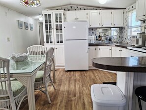 Cocina compacta con armarios blancos, un refrigerador blanco, una mesa de comedor con sillas de mimbre y una mesita redonda.