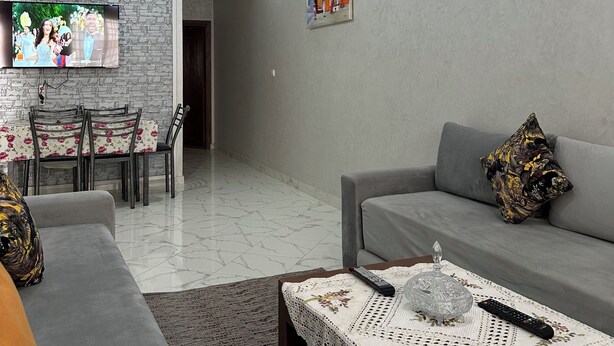 A modern living room with a grey sofa, a coffee table with a lace tablecloth, a TV mounted on a textured wall, and a dining area with a floral tablecloth. - Enjoy staying in a calm apatment- near the beach with air conditioning and wifi (Martil)