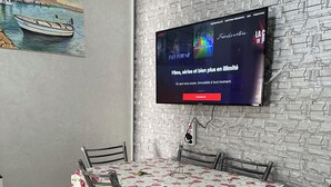 A dining area with a table covered in a floral tablecloth, surrounded by metal chairs. A flat-screen TV mounted on a textured wall displays a French film service. - Enjoy staying in a calm apatment- near the beach with air conditioning and wifi (Martil)