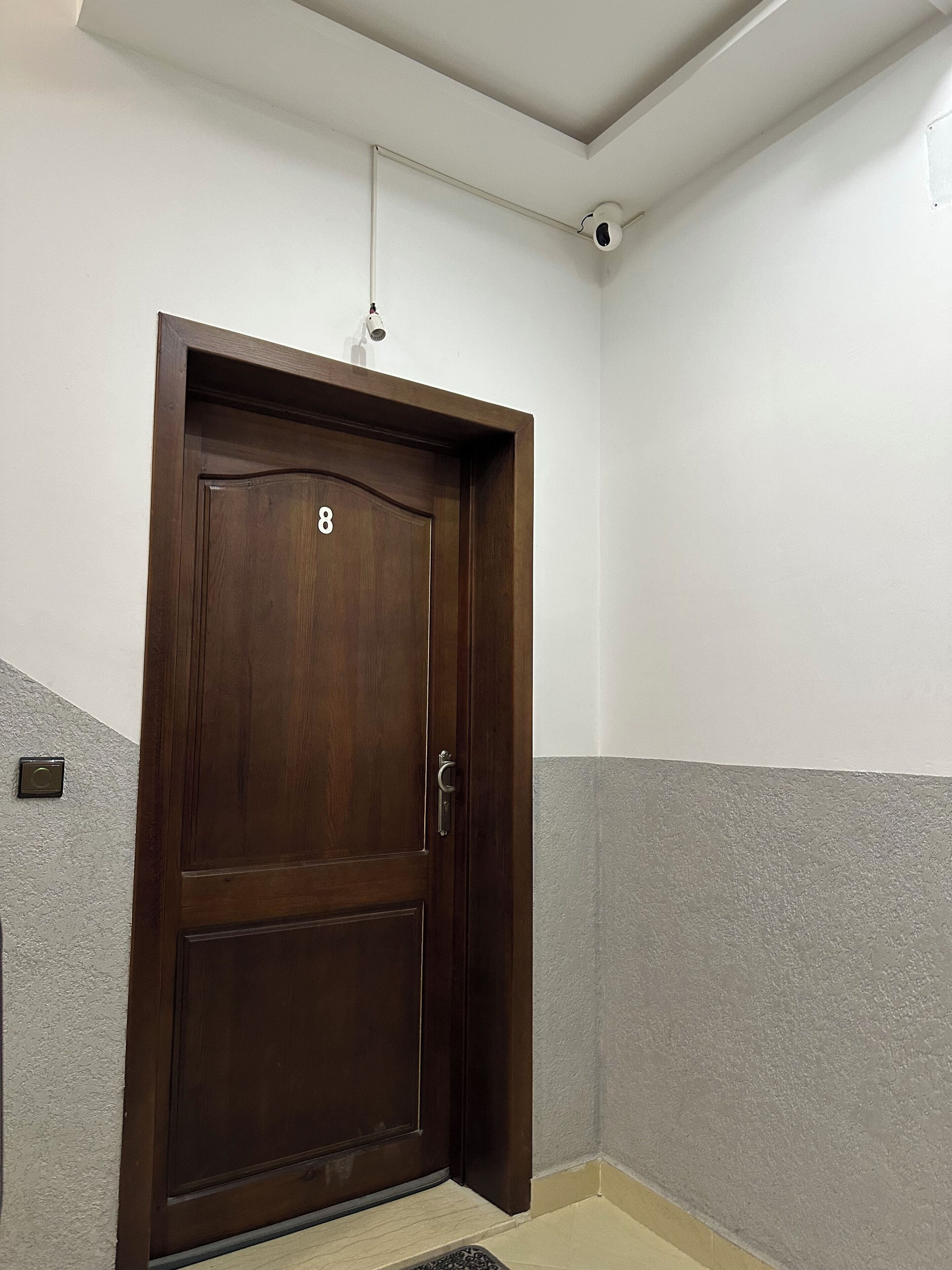 Uma porta de madeira escura com o número 8, um puxador de metal e uma câmera de segurança acima.