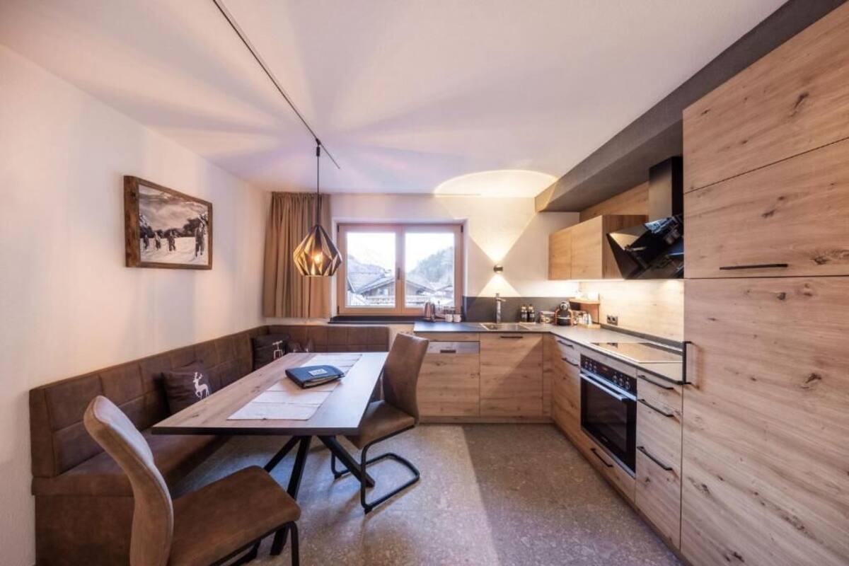 Een kleine keuken met houten kastjes, een eethoek met tafel en stoelen, en een raam met uitzicht op bergen.