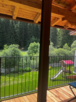 A wooden deck with a metal railing, a playground with a slide, and a forested background. - Wildbach Apartments Stubai (Neustift Im Stubaital)
