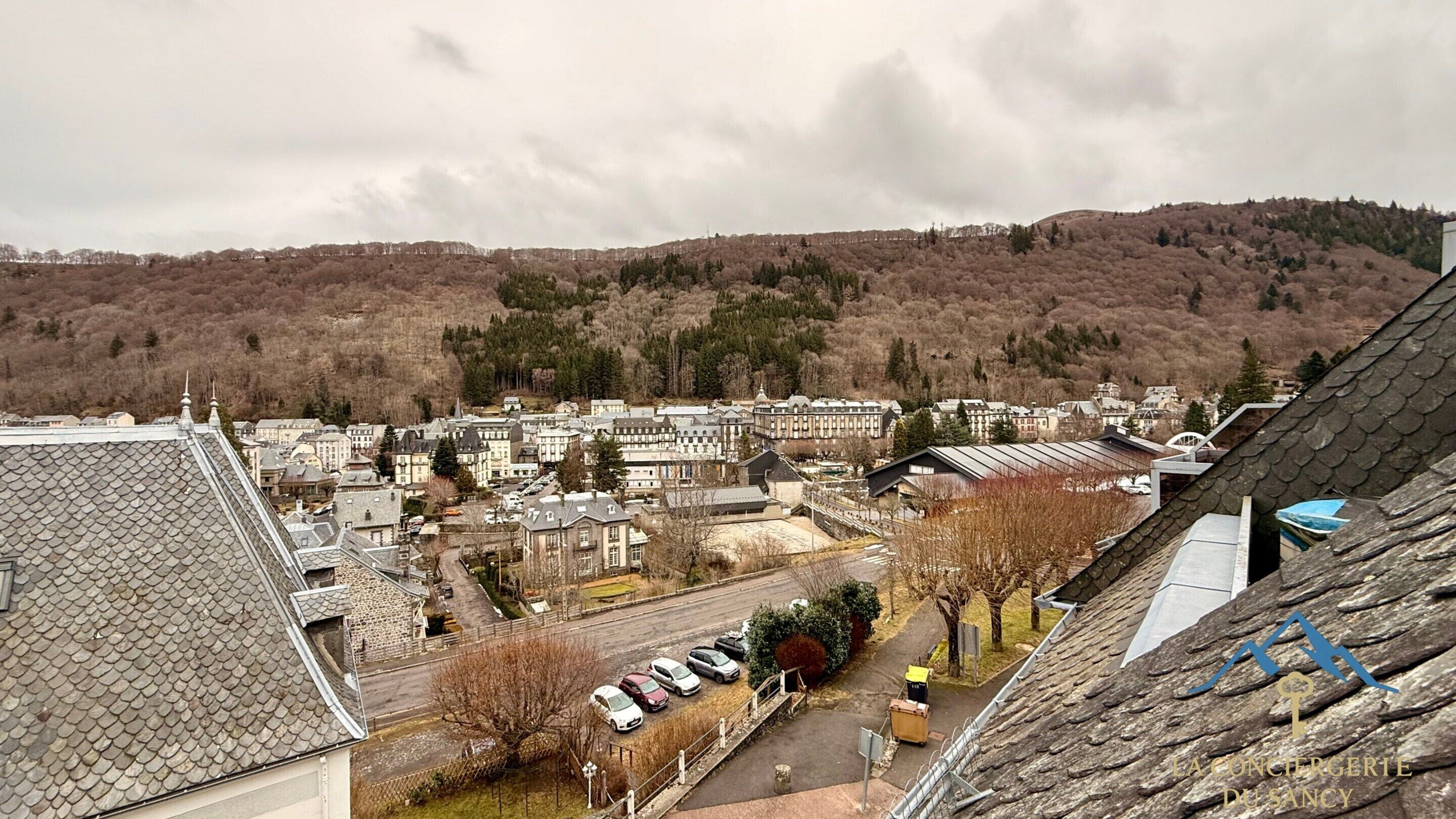 A town with a mix of residential and commercial buildings, a parking area, and a hilly landscape in the background.
