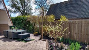 Een terras met een tafel en stoelen met een glazen blad, omgeven door een tegelpad en een houten hek.