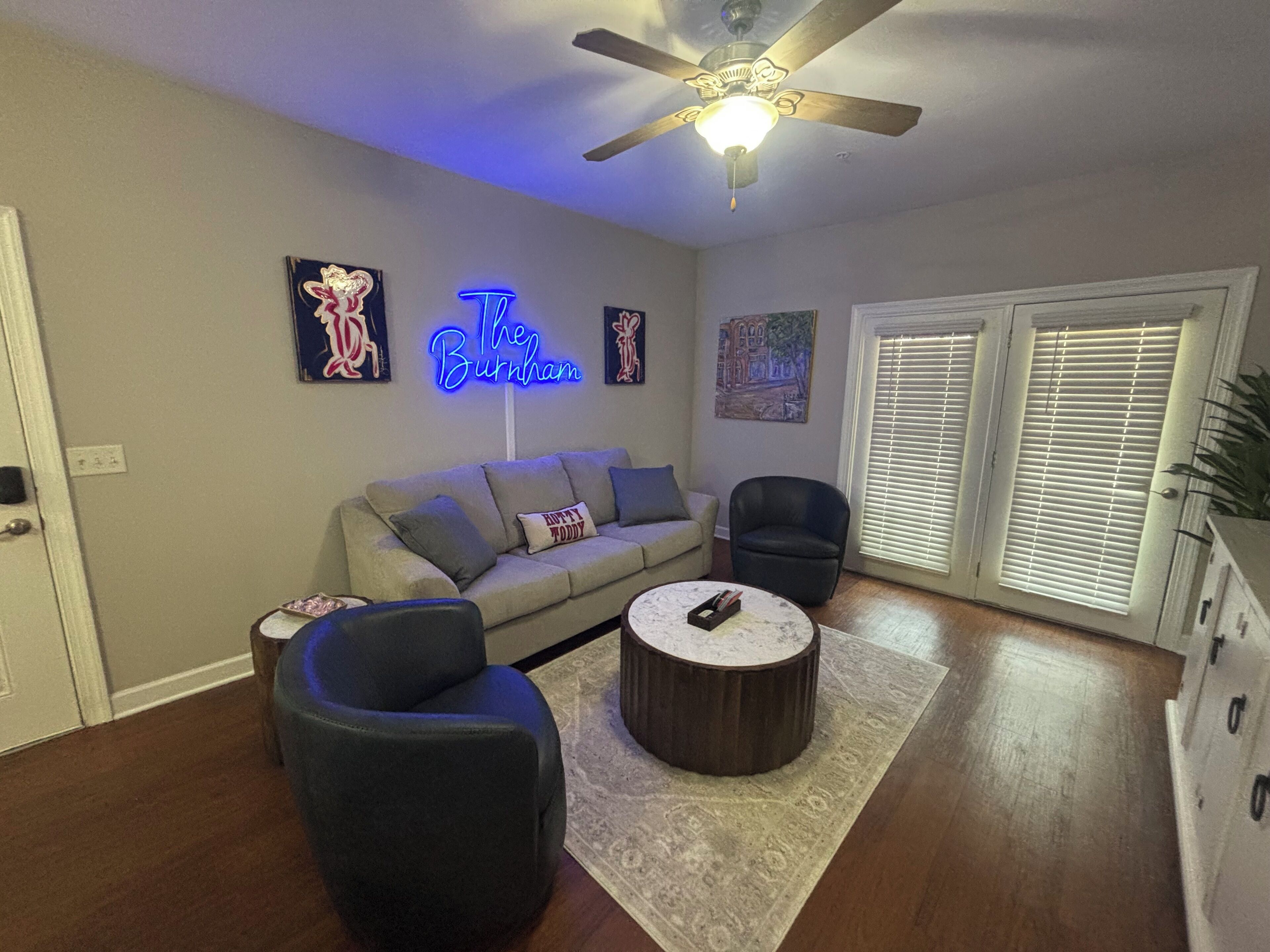A living room with a sofa, two armchairs, a coffee table, a ceiling fan, and a neon sign.