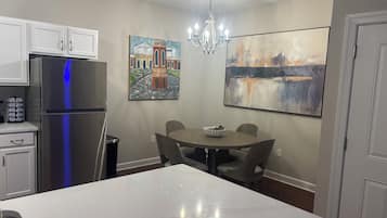 A modern kitchen with white cabinets, a stainless steel refrigerator, a chandelier, and two paintings on the walls.