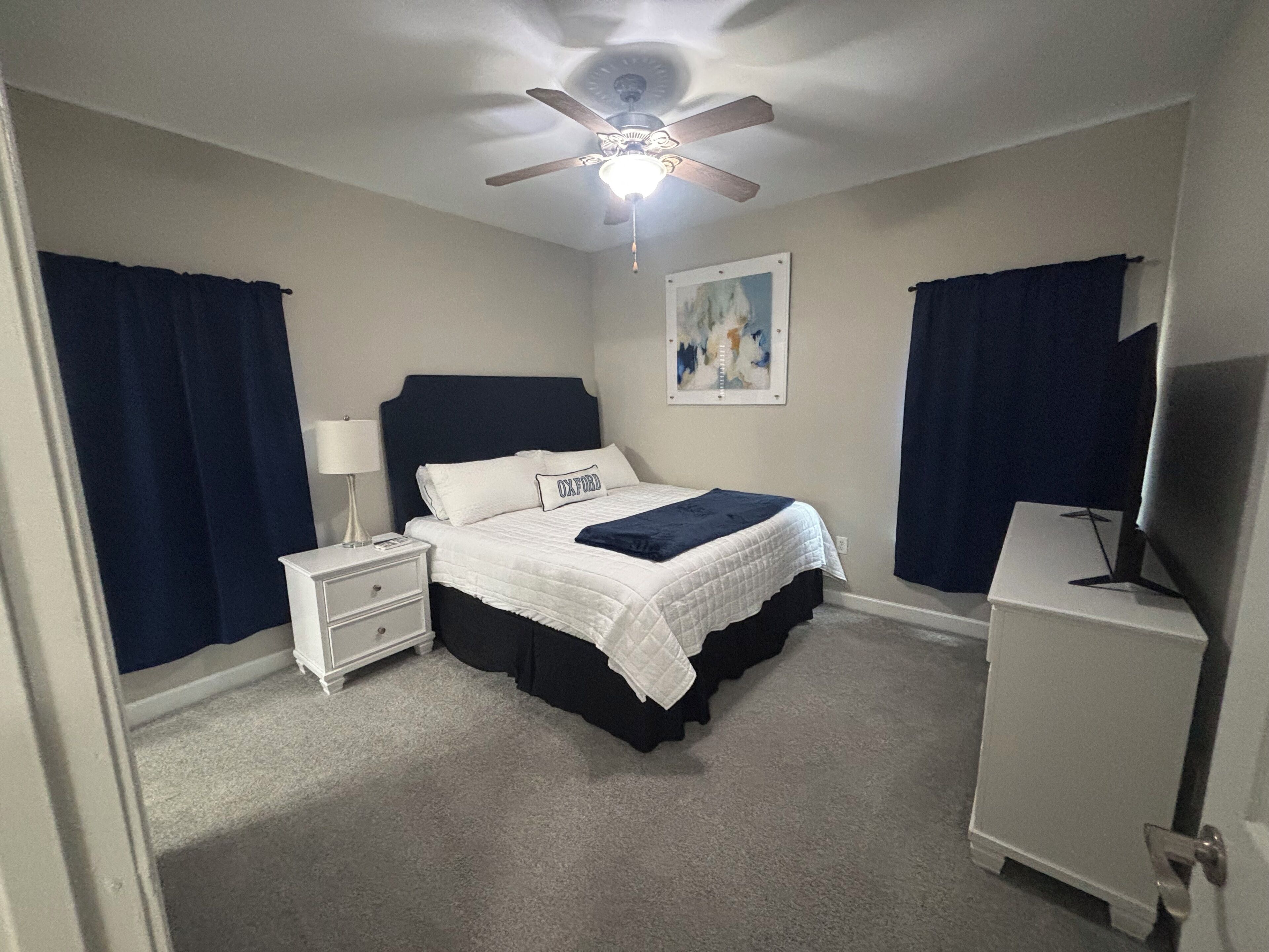 Ein Schlafzimmer mit Bett, Nachttisch, Lampe, Deckenventilator, Kunstwerk und einem Fernsehmöbel.