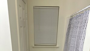 Un baño con una puerta blanca, una ventana con persianas, un perchero con tres toallas, una cortina de ducha y un felpudo negro.