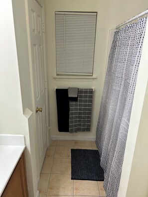 A bathroom with a white door, a window with blinds, a towel rack with three towels, a shower curtain, and a black doormat. - Relaxing Stay in Friendly Home (Charlotte)
