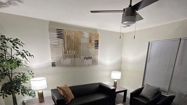 Una sala de estar moderna con un sofá de cuero negro, dos mesas auxiliares con lámparas, una mesa de centro, un ventilador de techo y una gran obra de arte abstracta en la pared.