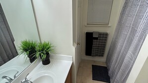 Un baño con lavamanos blanco, espejo, planta en maceta, toallero, cortina de ducha y ventana con persianas.