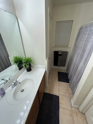 A bathroom with a white sink, a mirror, a potted plant, a towel rack, a shower curtain, and a window with blinds. - Relaxing Stay in Friendly Home (Charlotte)