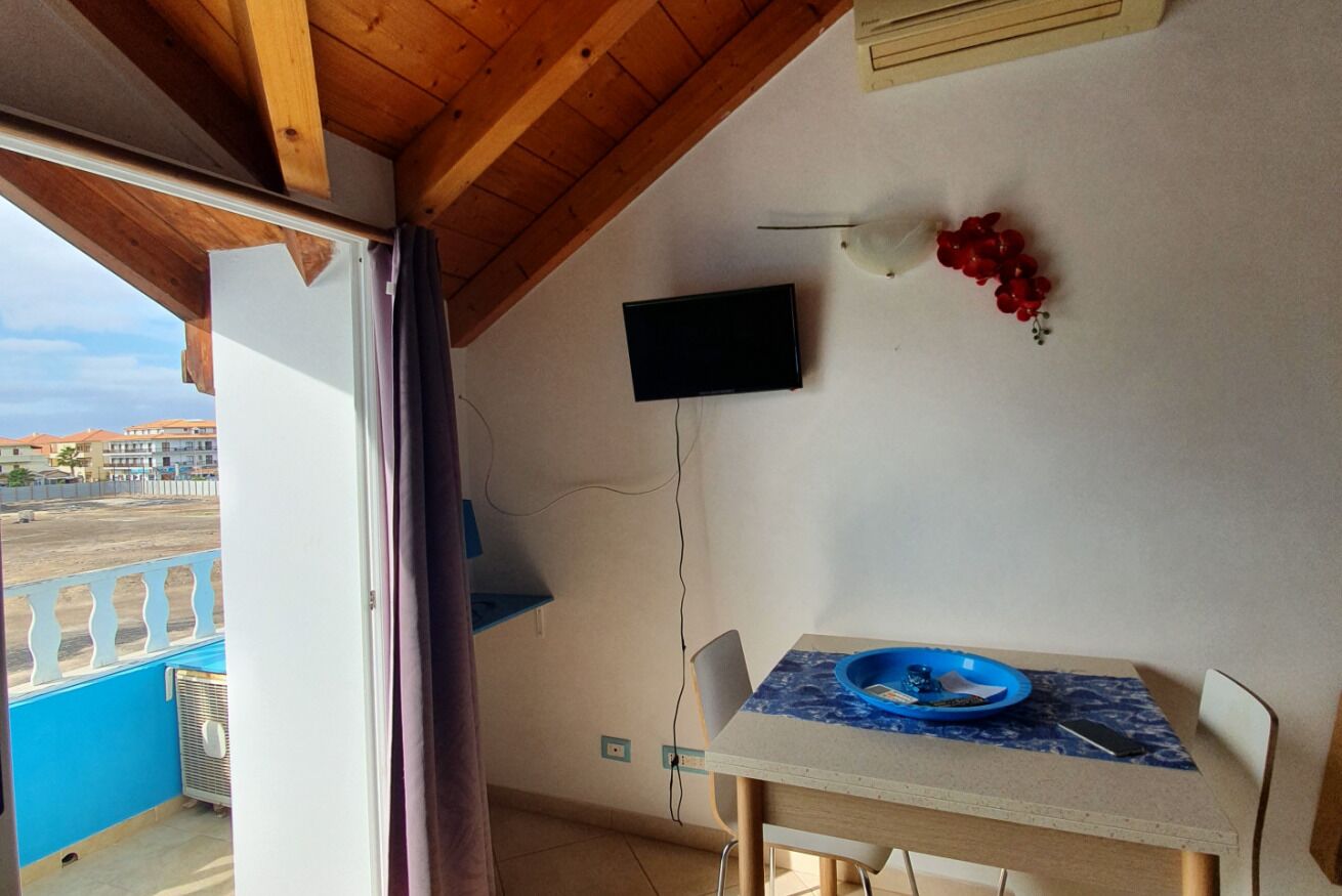 Een kamer met een houten plafond, een aan de muur bevestigde televisie, een eettafel met een blauw bord en uitzicht op buiten.