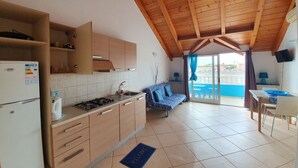 A compact kitchen with wooden cabinets, a gas stove, and a refrigerator. There's a blue sofa and a dining table with chairs. The room has a sloped wooden ceiling and a view through a glass door. - Holiday Properties Soleil (Santa Maria)