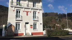 A two-story building with a white facade, red trim, and a large red 'T' on the front. There are balconies on the second floor, and a small porch with steps leading to the door. - Cozy French Alpine Retreat for 2 (Mont-Dore)