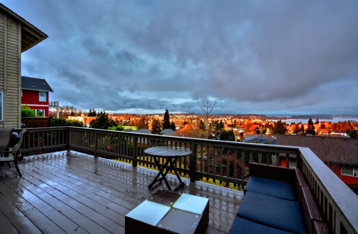 Um deck de madeira com mesa e cadeiras, com vista para uma paisagem urbana com árvores em tons de outono e céu nublado.