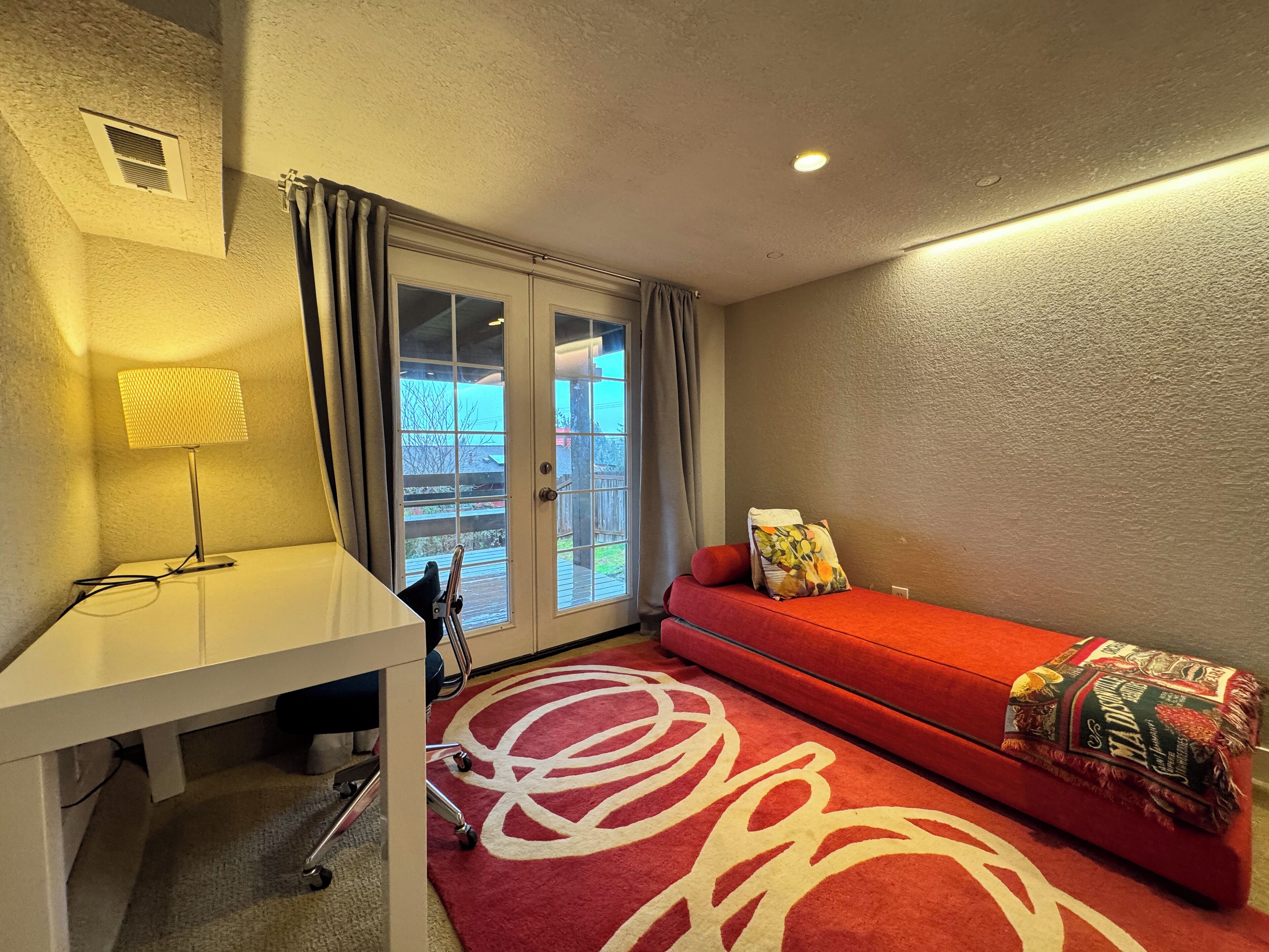 Une pièce avec un canapé rouge, une lampe, un bureau et une vue sur l’extérieur, visible à travers une porte vitrée.