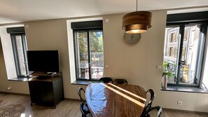 A modern living room with a large window, a flat-screen TV, and a dining table with chairs. - Au Coeur du Mont Dore : 6 Personnes (Mont-Dore)