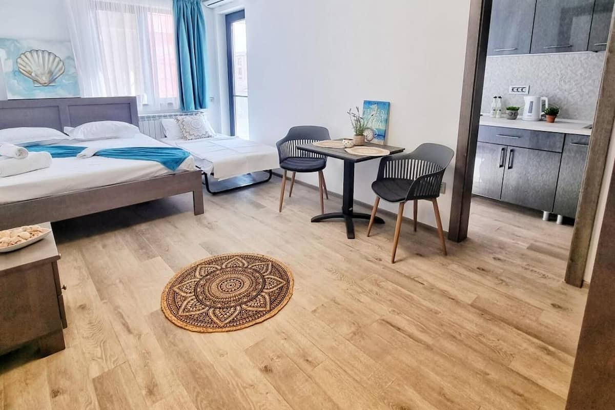 A modern, well-lit room with a bed, two chairs, a small table, and a kitchen area.