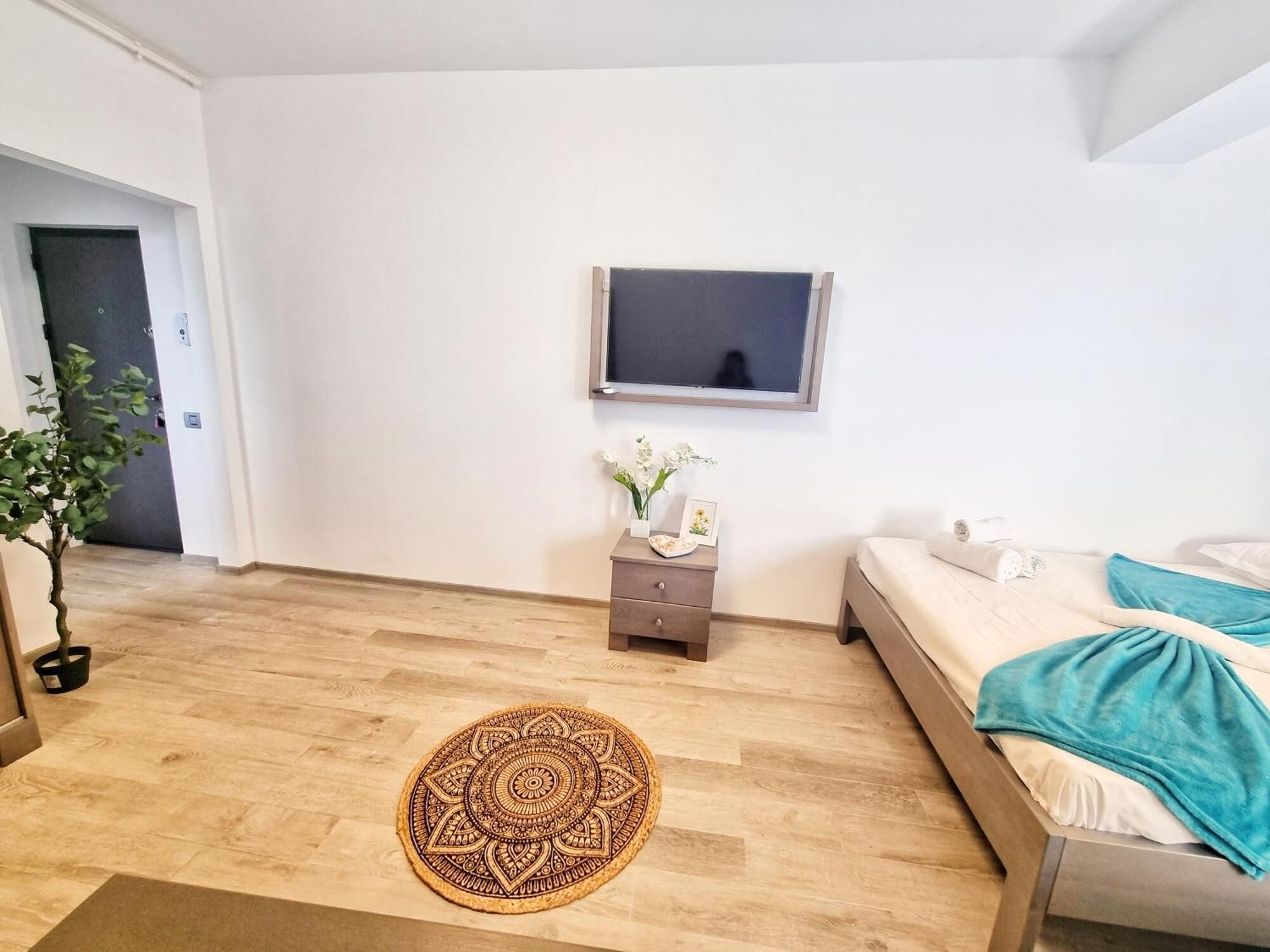 Un dormitorio moderno con cama, una mesita de noche con un florero, televisor montado en la pared y piso de madera.