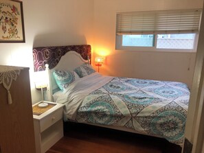 Un dormitorio con cama, mesita de noche, lámpara, ventana con persianas y un cuadro en la pared.