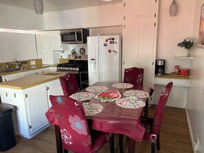 Cocina pequeña con armarios blancos, microondas, estufa y refrigerador. Hay una mesa de comedor con posavasos estampados y sillas rojas.