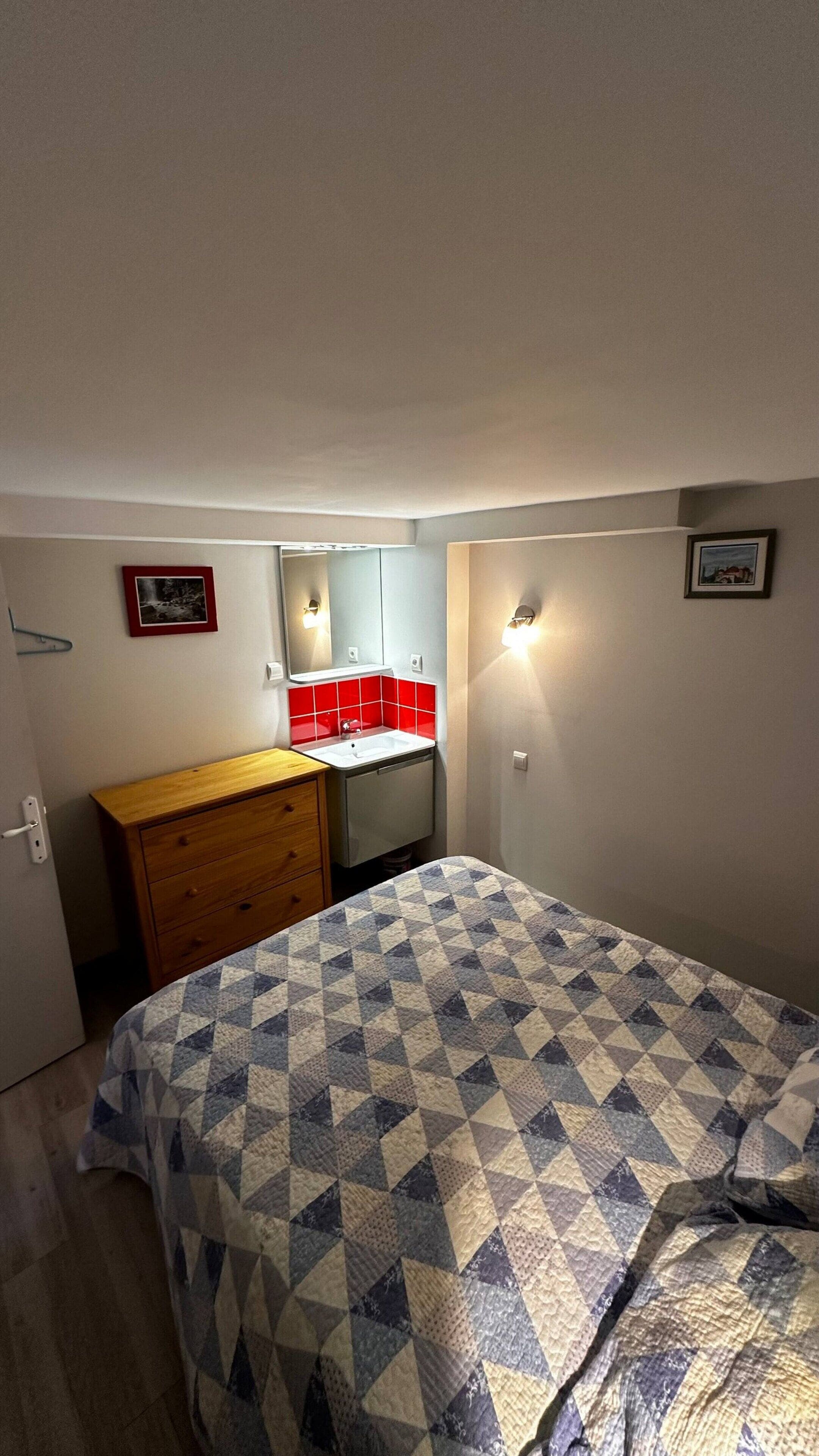 Une petite chambre à coucher, bien rangée, avec un lit, une commode en bois, un lavabo aux carreaux rouges et un miroir.