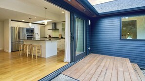 Une cuisine moderne dotée d’un îlot central, de tabourets en bois et d’une porte-fenêtre coulissante donnant sur une terrasse.