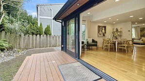 Une maison moderne dotée d’une terrasse en bois, d’un coin repas avec une table ronde et des chaises, et d’une grande porte-fenêtre coulissante donnant sur l’intérieur.