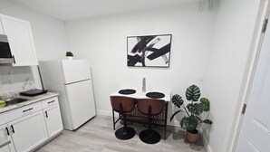 Een kleine keuken met witte kastjes, een witte koelkast, een eethoek met twee stoelen en een tafel, en een grote plant.