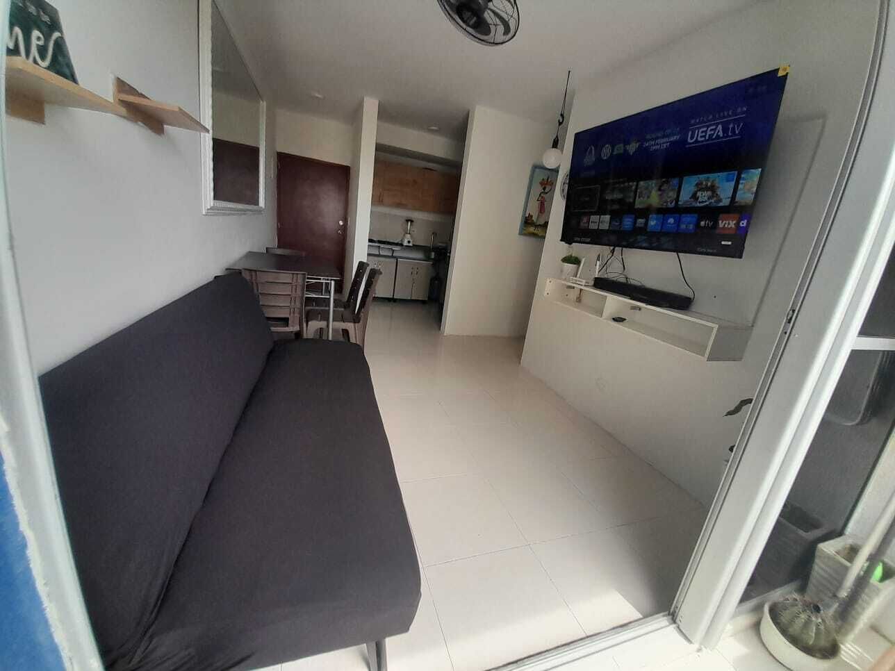 A compact living space with a sofa, a TV mounted on the wall, and a kitchenette in the background.