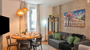 Uma sala de estar moderna com uma mesa de jantar redonda de madeira, sofá cinza com almofadas verdes e um quadro de um edifício na parede.
