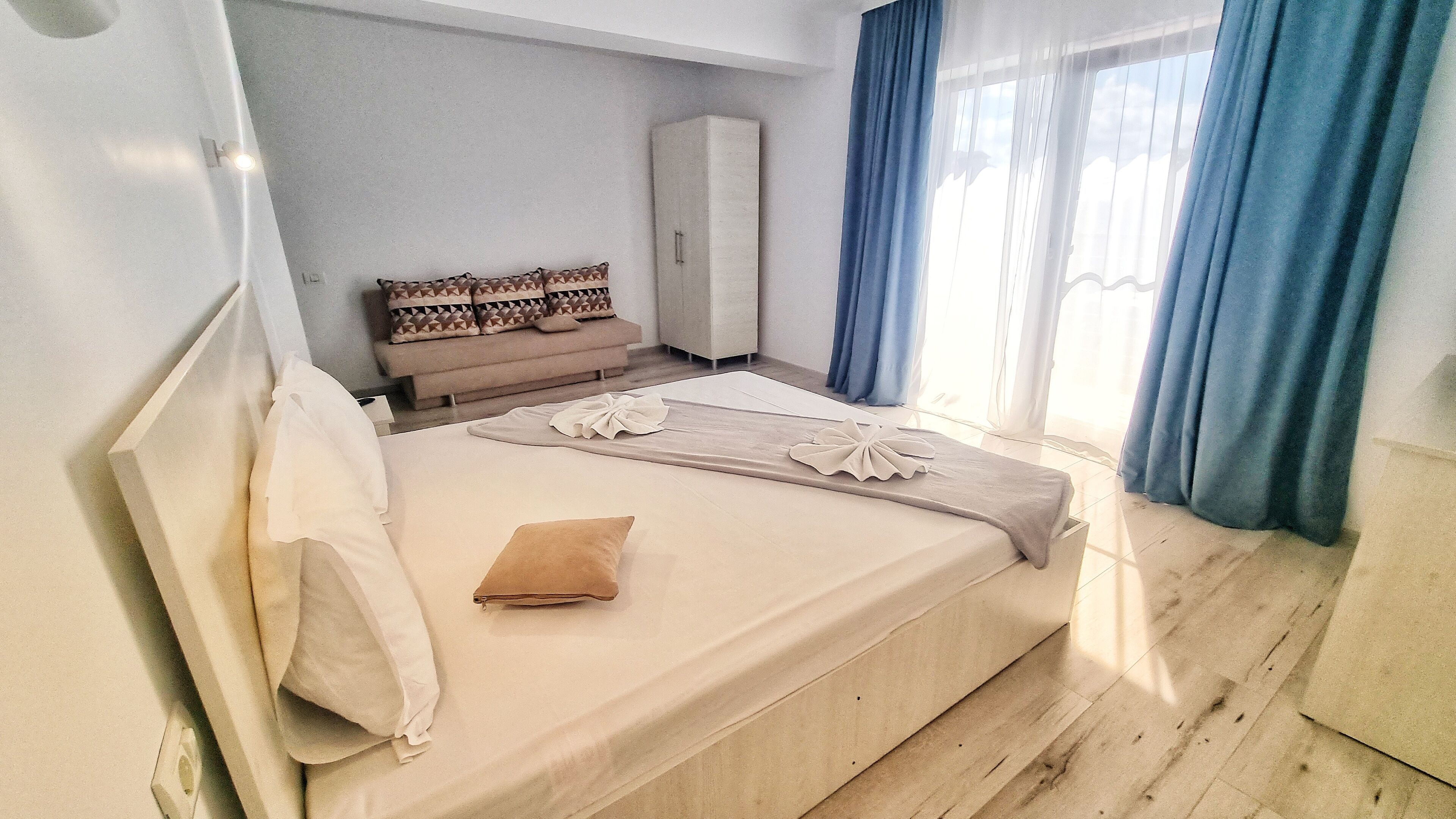 Una cama bien hecha con una almohada y una toalla doblada, un armario, un sofá con cojines estampados y una ventana con cortinas azules.