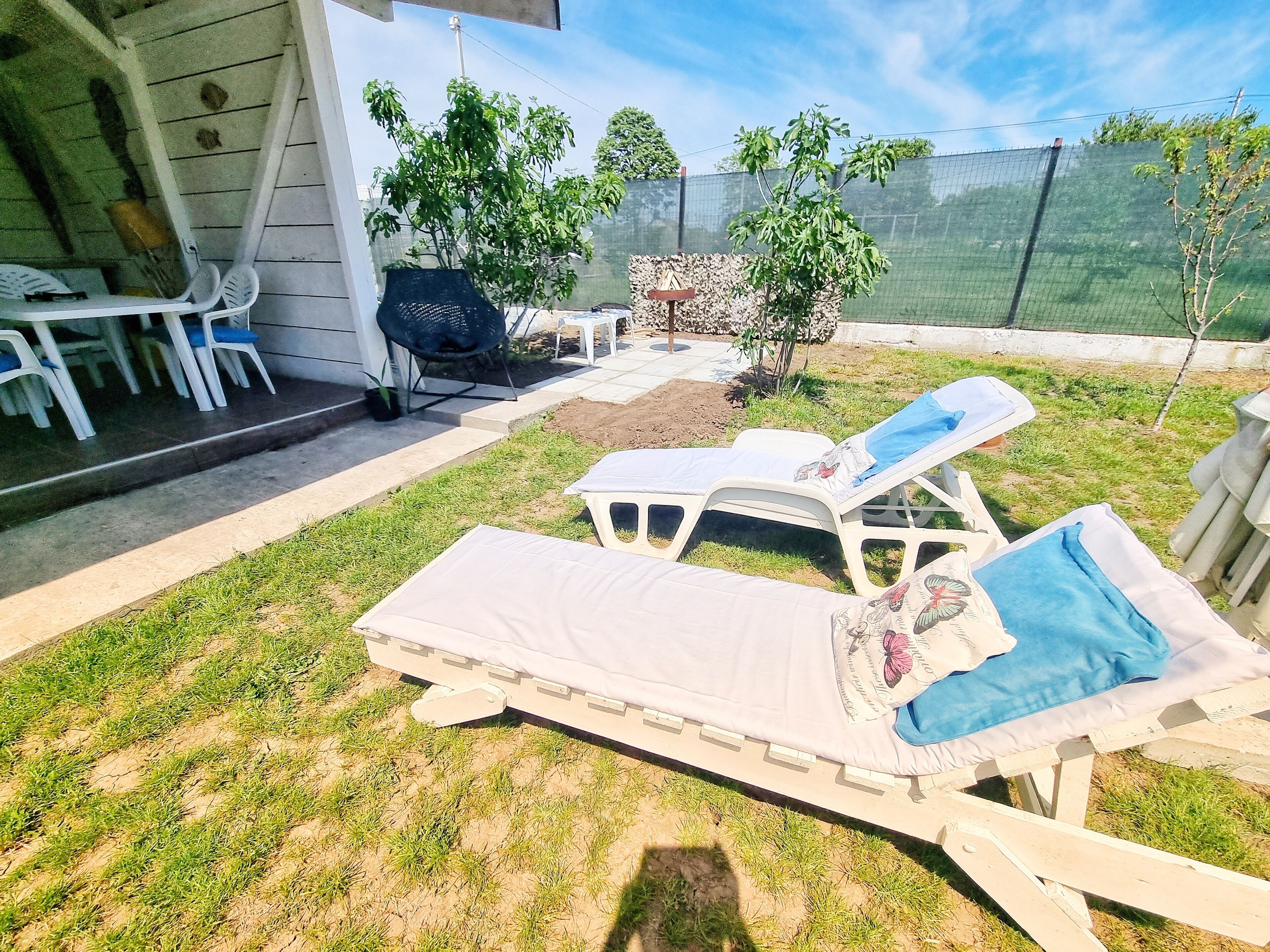 Un espace patio aménagé avec deux chaises longues, une petite table et une chaise. On aperçoit des plantes en pot et une clôture à l’arrière-plan.
