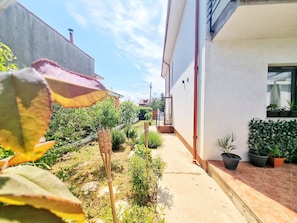 A narrow pathway leading to a building with a white exterior and a red roof. There are potted plants along the pathway and a small garden area with various shrubs and a bamboo structure.