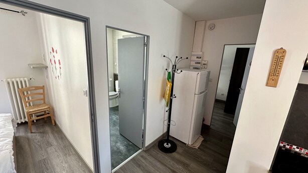 A compact room with a wooden chair, a radiator, a small shelf, a mirror, a coat rack, a refrigerator, and a door leading to another room. - Mountain View Flat for 5 in Mont-dore (Mont-Dore)