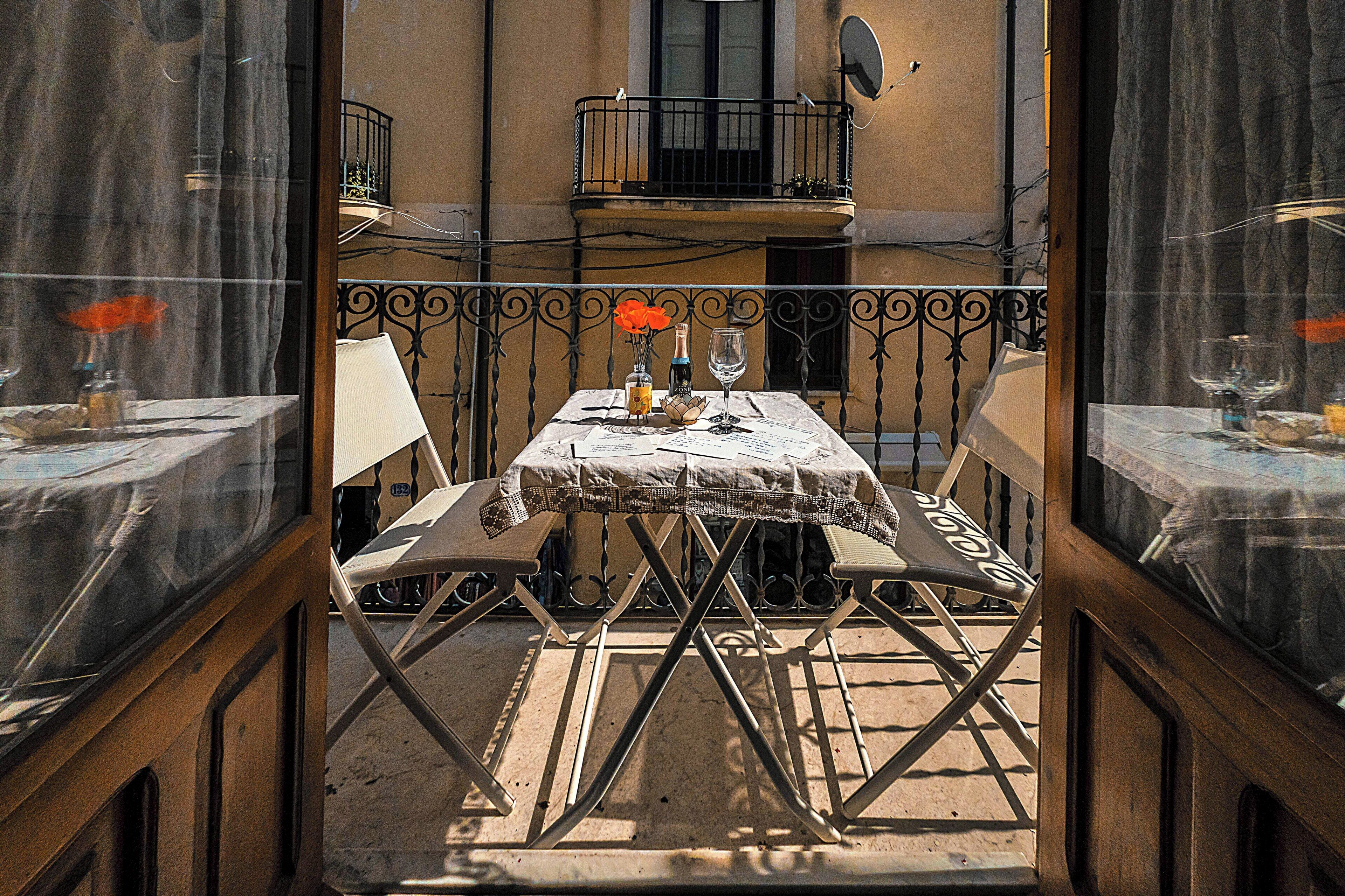 An outdoor dining area with a table set for two, a balcony with a railing, and a building in the background.