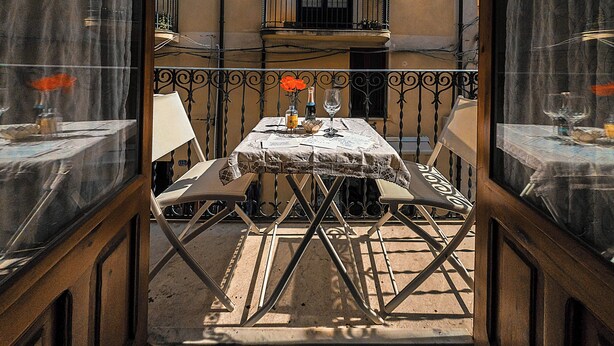 An outdoor dining area with a table set for two, a balcony with a railing, and a building in the background. - Angelina's apartment (Cefalù)