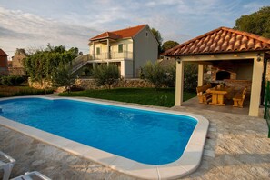 A swimming pool with a stone deck, a covered outdoor kitchen, and a two-story house with a red-tiled roof. - Villa in a Sunny Trbounje Zone (Drnis)