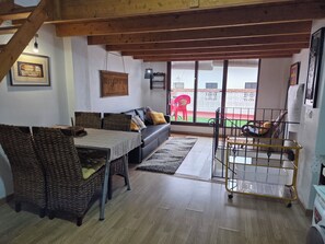 Un salon comprenant un canapé, une table à manger, des chaises en osier, un chariot en métal et une vue sur un espace extérieur, accessible par une porte vitrée.